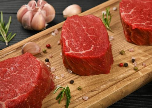 Close up three aged prime marbled raw tenderloin or fillet mignon beef steaks on brown oak wood cutting board, with spices, over black wooden table background, high angle view