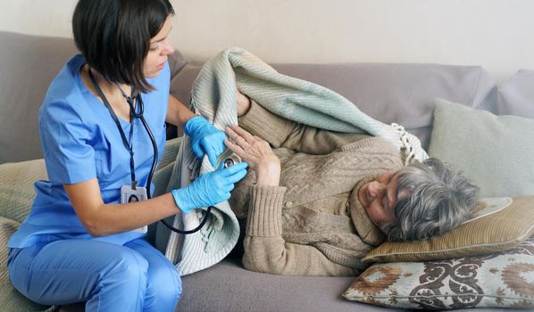 A young nurse is caring for an elderly 80-year-old woman at home, she asks about the well-being of a pensioner who lies and rests in bed. Happy retired woman and trust between doctor and patient.