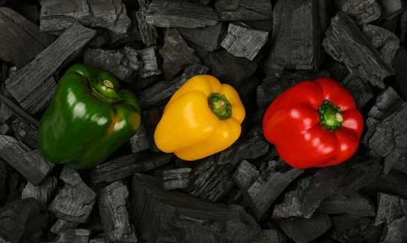 Close up raw red, yellow and green bell peppers on black lump charcoal pieces ready for barbecue grill cooking, elevated top view, directly above