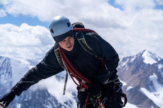 A young man traveler is engaged in mountaineering.In a helmet, with a rope, a harness, gloves, climbs to the top, against the backdrop of a stunning view with snow-capped mountains.