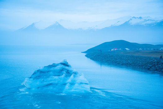 Icelandic glacier floating from global warming effects. Foggy atmosphere with snowcapped mountain range in the distance. Iceberg has layers of volcanic ash.