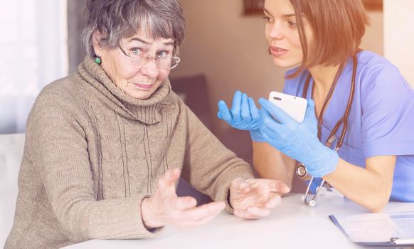 A nurse in a medical suit takes care and explains to an elderly patient how to use applications on a smartphone. Grandmother 80 years old, does not understand how to communicate with a doctor online.