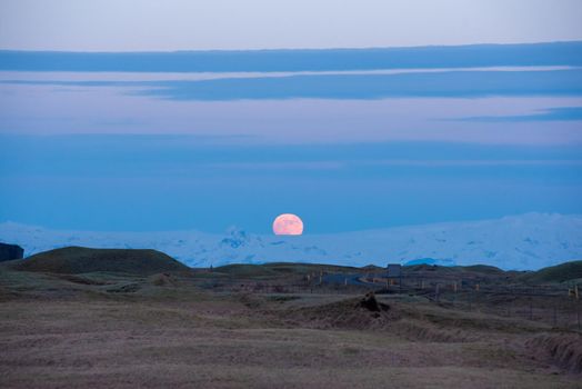 Full moon rise over Iceland glacier with rolling hills in the foreground and deep blue sky