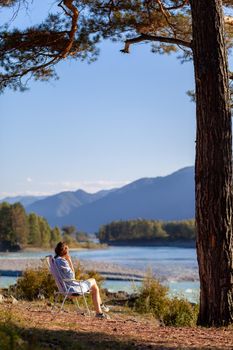 A woman is sitting on a folding chair on the bank of a mountain river on a nice, warm day under a large tree. A calm and quiet place to relax and reflect. Equipment and a tourist's rest.