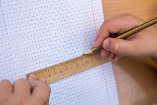 The student draws a figure in a notebook with a pencil along a ruler. A schoolboy performs a task at the workplace. The concept of children's education, teaching knowledge, skills and abilities