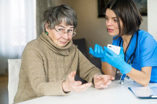 A nurse in a medical suit takes care and explains to an elderly patient how to use applications on a smartphone. Grandmother 80 years old, does not understand how to communicate with a doctor online.