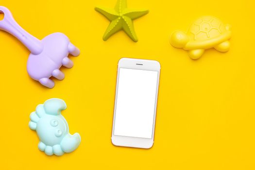 Mobile phone with white screen and plastic beach toy pastel color on yellow background. The development of fine motor concept. Creativity Game, technology and summer concept. top view