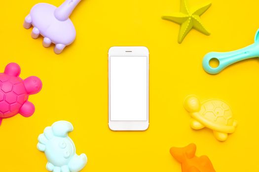Mobile phone with white screen and plastic beach toy pastel color on yellow background. The development of fine motor concept. Creativity Game, technology and summer concept. top view