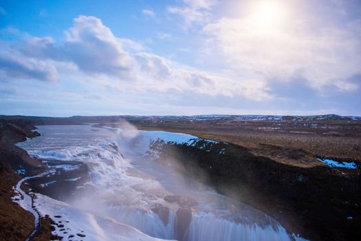 Godafoss waterfall in Iceland at sunset with atmospheric landscape including mist and puffy clouds in blue sky with sun