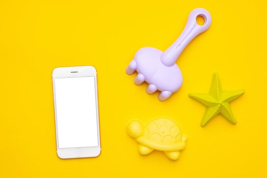 Mobile phone with white screen and plastic beach toy pastel color on yellow background. The development of fine motor concept. Creativity Game, technology and summer concept. top view