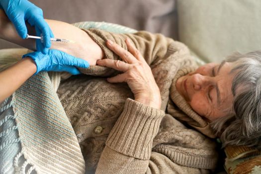 Nurse makes an injection with the vaccine to the patient at home. A young doctor in a blue suit holds the hand of an elderly 80 age woman and makes injection with a syringe. Medicine and healthcare.