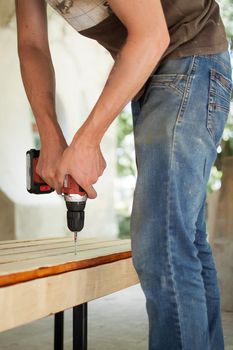 Skilled young male worker is using power screwdriver drilling during construction wooden bench, do it yourself.