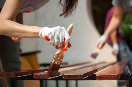 Female worker makes painting works of wooden products, plank with brown paint for making bench, carpenters work, wood covering protection