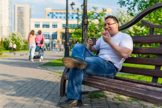 Happy man enjoying delicious donut and drink, laughing while sitting on a park bench