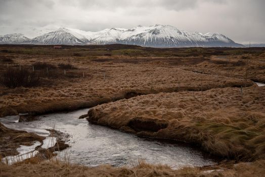 Icelandic landscape with snaking stream through grassy field with snow capped mountains in the distance atmospheric cloudy textured