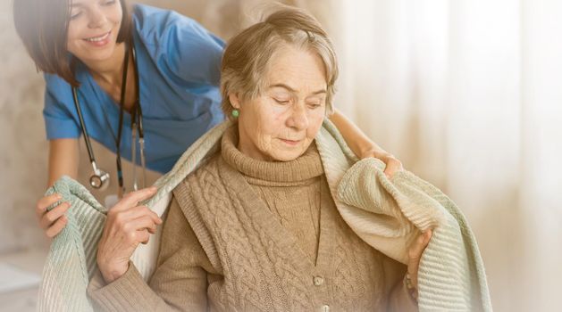 A young nurse takes care of an elderly 80 year-old woman at home, wraps a blanket around her. Happy retired woman and trust between doctor and patient. Medicine and healthcare.