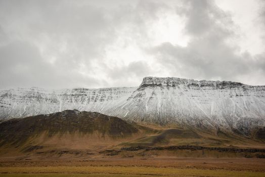 Heavenly light spills through the clouds over a snow capped mountain ridge leading down into brown grassy landscape in Iceland