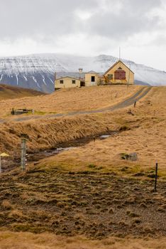 Grassy farmland with driveway leads to a home with a blue mountain snowy backdrop in Iceland