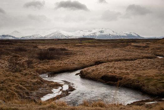Icelandic landscape with snaking stream through grassy field with snow capped mountains in the distance atmospheric cloudy textured