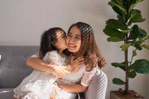 Sweet little girl is hugging and kissing her beautiful young mom in cheek while sitting on couch at home