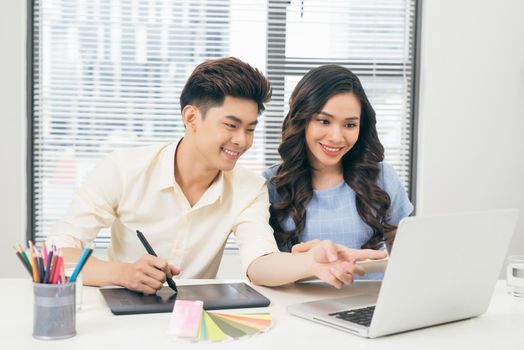 Two smiling casual designers working with laptop while sitting at desk in the office