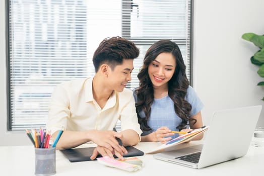 Two smiling casual designers working with laptop while sitting at desk in the office