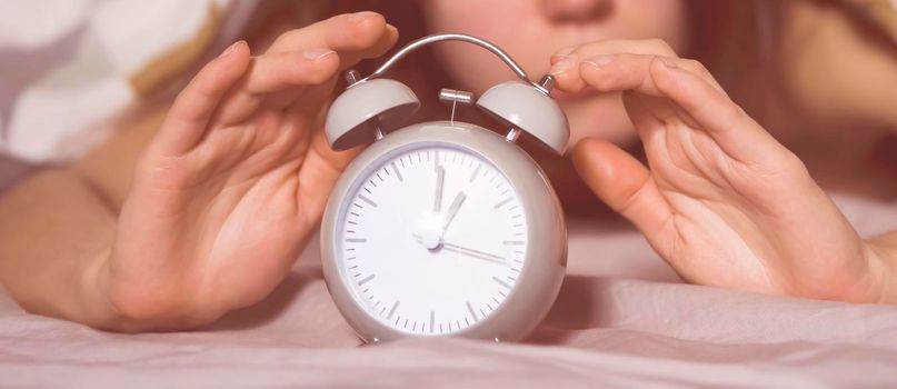 Hands of a young girl from under the blankets hold a retro vintage alarm clock in gray. The person holds a clock, need to wake up.