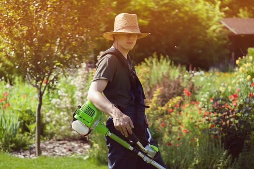 A young man in a straw hat is mowing a lawn with a lawn mower in his beautiful green floral summer garden. A professional gardener with a lawnmower cares for the grass in the backyard.