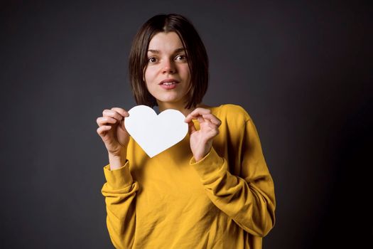 Young girl holds a white paper heart - a postcard in order to write about love. A woman with a joyful surprised face in anticipation of the holiday and in search of love and romantic relationships.