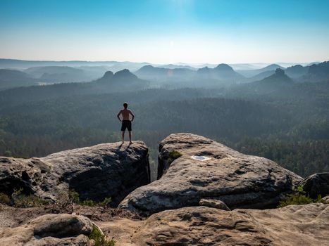 Tall shirtless male sporty figure on the edge of a rock enjoys the view of the morning landscape.