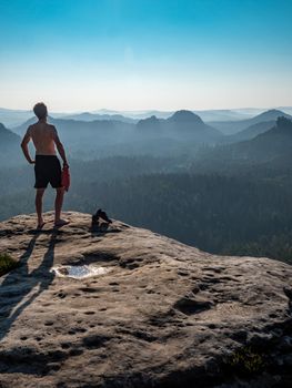 Shirtless man with a sporty figure on the edge of a rock enjoys the view of the morning landscape.