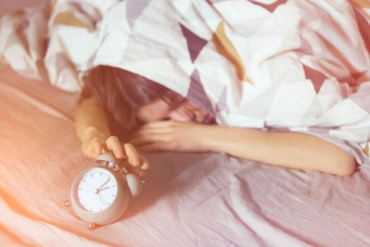 A young girl sleeps early in the morning under the blanket and holds a vintage alarm clock in her hands.