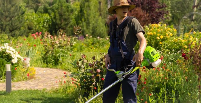 A young man in a straw hat is mowing a lawn with a lawn mower in his beautiful green floral summer garden. A professional gardener with a lawnmower cares for the grass in the backyard.