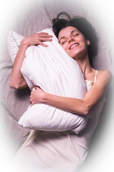 A happy, cute girl with a smile on her face gently hugs a white soft pillow, lies in bed and feels comfortable. Young woman wakes up in a cozy home, holding a pillow in her hands.