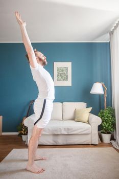Mid adult mature man in white sport clothes practice yoga stretch doing exercise at home minimalistic scandinavian design room