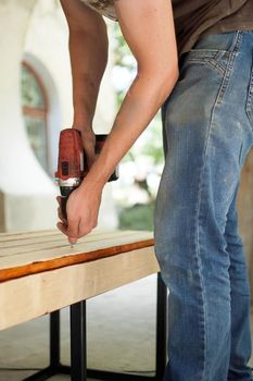 Skilled young male worker is using power screwdriver drilling during construction wooden bench, do it yourself.