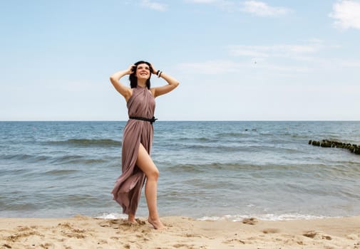 young brunette woman in beige dress stands on the sand by the sea on summer day