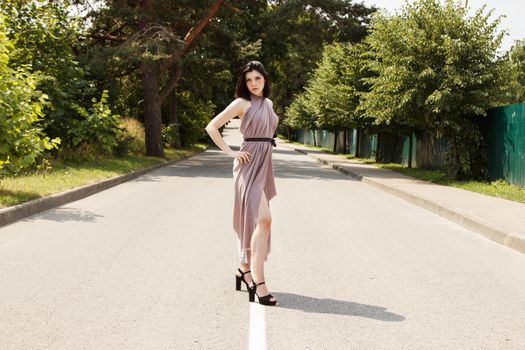 young beautiful brunette woman in beige dress standing on the road in summer