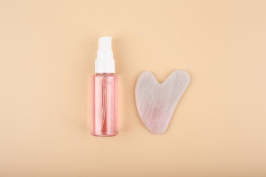 Flat lay with massage gel, skin serum or oil in transparent glass bottle and heart shaped guasha pink quartz crystal on bright beige background. Concept of facial massage and self treatment