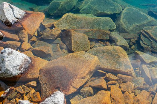 Beautiful coastal stones of turquoise and orange colors, partially lying under the clear fresh water of the lake