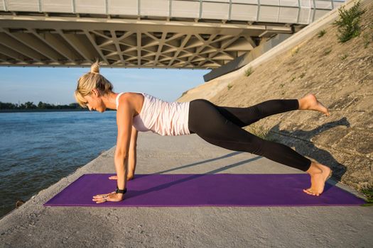 Young woman is exercising outdoor. She is practicing pilates. Plank with leg lift exercise.