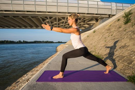 Young woman is exercising outdoor. She is practicing yoga on sunny day. Virabhadrasana / Variation of Warrior pose.