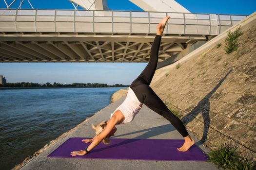 Young woman is exercising outdoor. She is practicing yoga on sunny day. Eka Pada Adho Mukha Svanasana/One-Legged Downward-Facing Dog Pose.