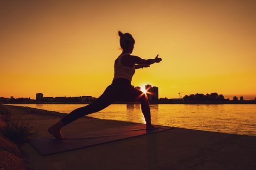 Young woman is exercising outdoor. She is practicing yoga on sunny day. Virabhadrasana / Variation of Warrior pose.