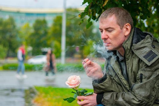 A smoking man with a rose sits in a park and thinks about something in the rain in the fall