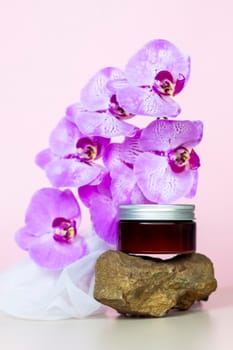 A brown jar of cream on a pink background stands on a large building stone with beautiful orchids next to it. Stylish appearance of the product, layout, personality. Banner, a place for the text.