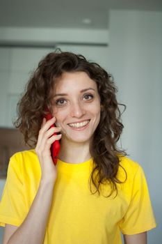 Beautiful curly girl in yellow t-shirt is talking with someone using red smartphone at home in her apartment, posing with smile, happy people concept.