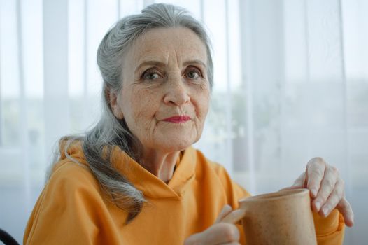 Happy dreamy middle aged woman sitting with cup of black tea or coffee, looking at camera. Peaceful mature lady enjoying no stress calm positive pastime alone at home. Happy retirement.