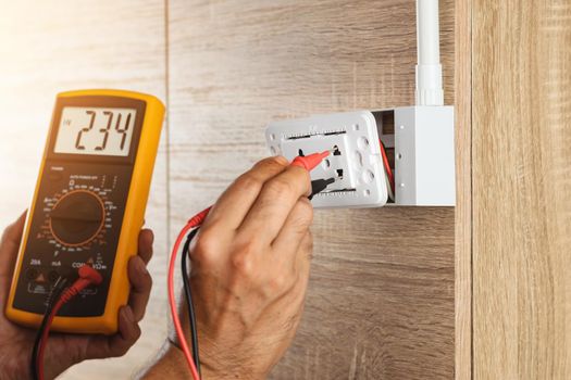 Electrician using a digital meter to measure the voltage at a wall socket on a wooden wall.
