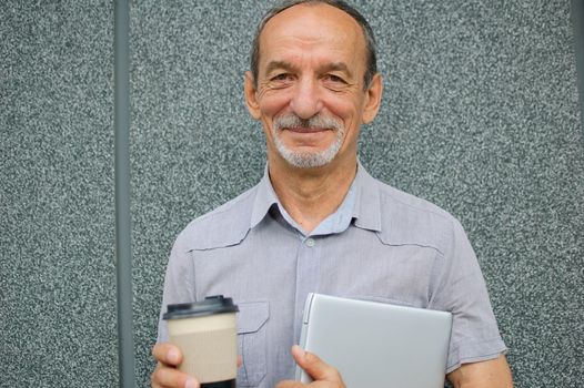 Male portrait of older coworker with silver laptop and cup of coffee waiting for a meeting near dark grey wall in the morning, remote job, coffee break.